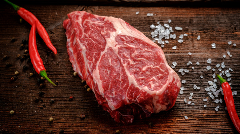 Raw chuck eye steak on cutting board alongside salt and red peppers