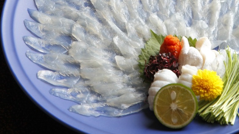 A light blue platter of thinly sliced blowfish intricately placed in a fan pattern with lime, herbs, and other accents on the side