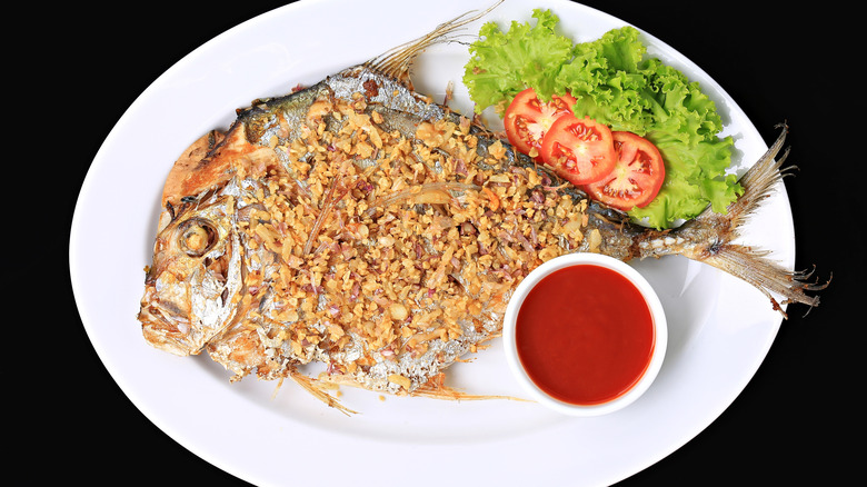 Trevally fish served with a side of tomato and lettuce on a plate next to a bowl of red dipping sauce