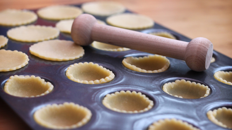 A muffin tray has dough being fitted into each cup with a special wooden tool.