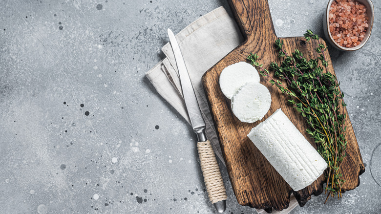A log of goat cheese is sliced into rounds on a cutting board beside a bunch of herbs.
