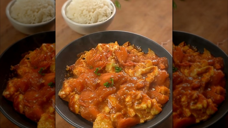 A saucy tomato egg stir-fry dish in a black plate with a bowl of white rice in the background