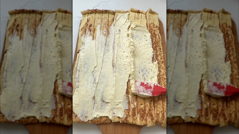 Person spreading cheesecake filling on unrolled canned cinnamon rolls