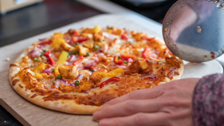 A persons hands slice up a pizza on a cutting board with a pizza wheel.