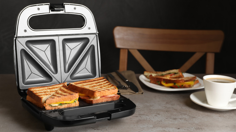 toastie grill maker with tasty sandwiches and cup of coffee on light grey table