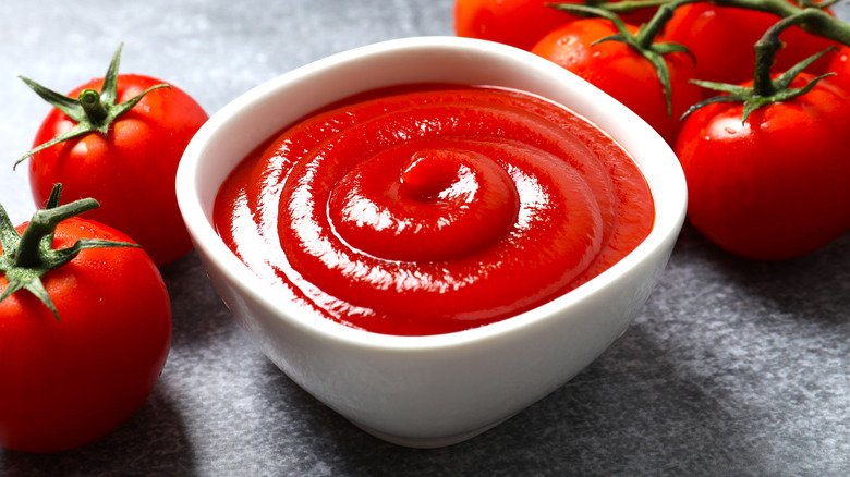 Ramekin of ketchup on a gray slate table surrounded by tomatoes