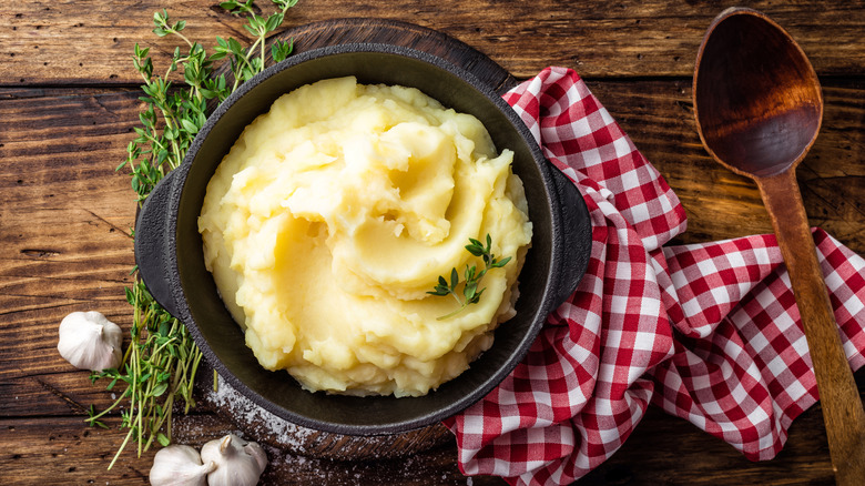 A pot of mashed potatoes is styled with herbs, a red-and-white gingham napkin, and a wooden spoon on a wooden surface