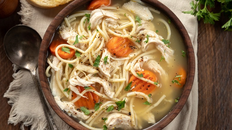 Bowl of chicken noodle soup with carrots on a napkin and baguette slices on a wooden table.