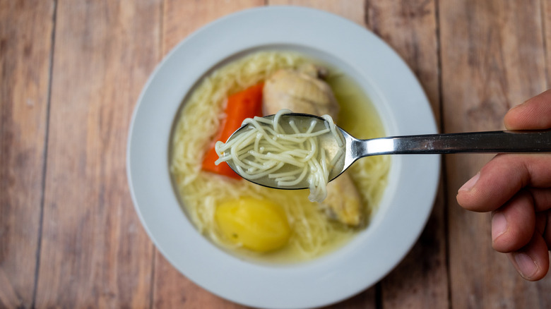 Top view of a traditional homemade chicken soup with noodles, vegetables, and chicken leg served in a bowl on a rustic wooden surface.