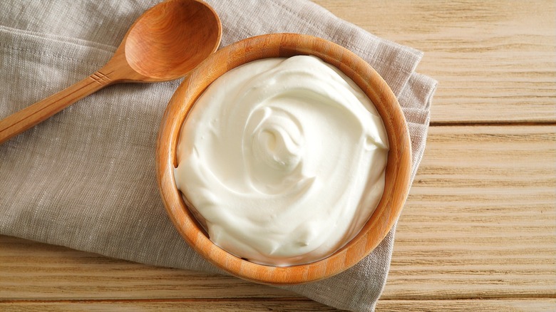 Greek yogurt in a small wooden bowl next to a wooden spoon over a folded cloth napkin.
