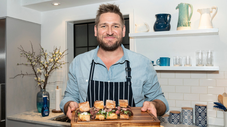 Curtis Stone in kitchen holding a board with sandwiches