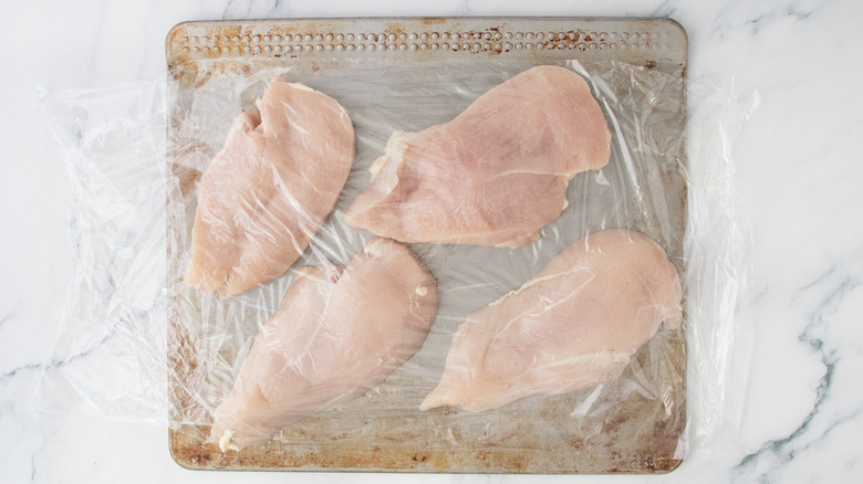 Chicken breasts covered in plastic wrap on a baking sheet.