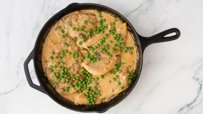 Seared chicken nestled into a cream sauce in a large skillet sprinkled with green peas