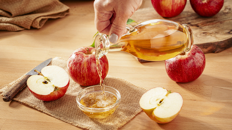 A person's hand is seen pouring apple cider vinegar into a small bowl with apples arranged in the frame.