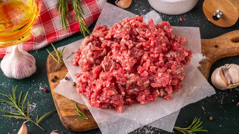 Ground beef rests on butcher paper on a cutting board surrounded by other ingredients like garlic and oil.
