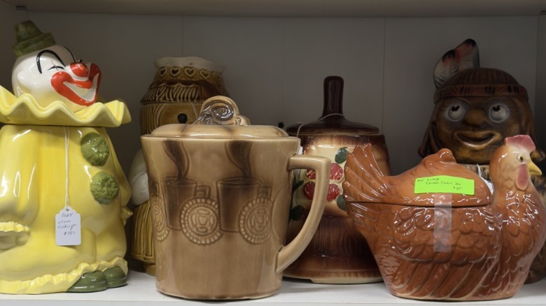Different kinds of vintage cookie jars lined up on a white shelf with price tags on them.