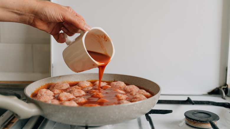 Hand pouring tomato sauce over meatballs in a silver skillet