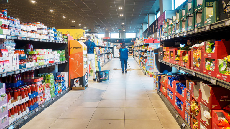 Customers and employees are walking through the aisles of a brightly lit ALDI supermarket, browsing the shelves stocked with various food products