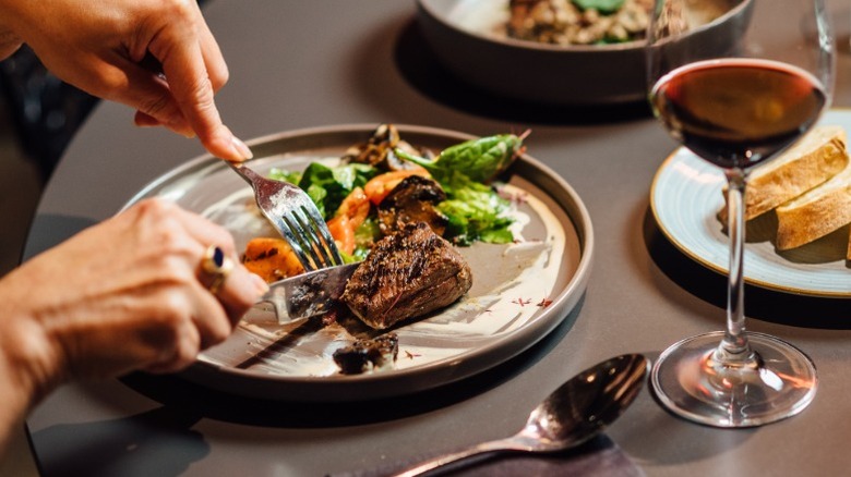 A restaurant customer cutting steak with a knife and fork, with other dinner items on the table surrounding them.
