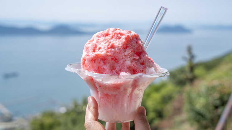 A hand holds a cup of pink shaved ice topped with condensed milk with a plastic spoon