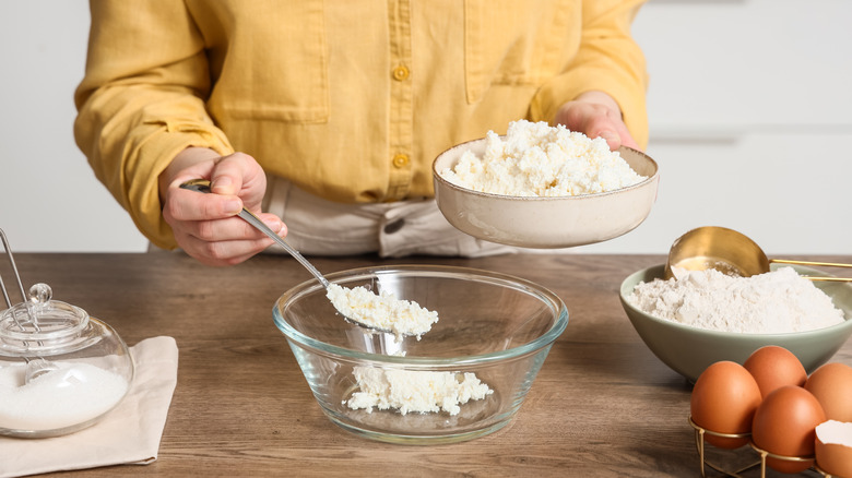 Person spooning cottage cheese into a bowl surrounded by cooking ingredients.