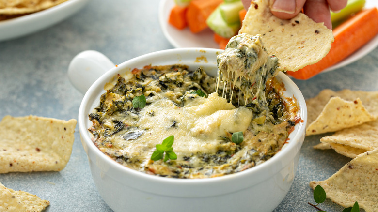 Hand dipping a tortilla chip in a dish of cheesy spinach dip.