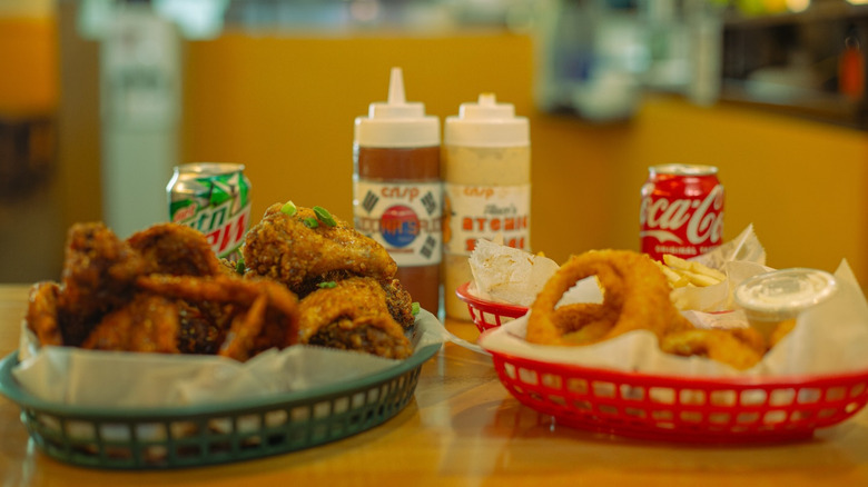 A basket of Korean fried chicken and onion rings with sauces and drinks in the background