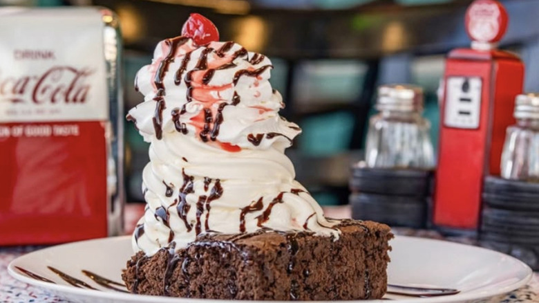 A brownie piled high with whipped cream, chocolate syrup, and a maraschino cherry