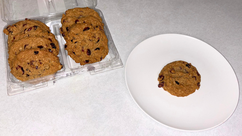 Container of cranberry oat cookies and one on a white plate.