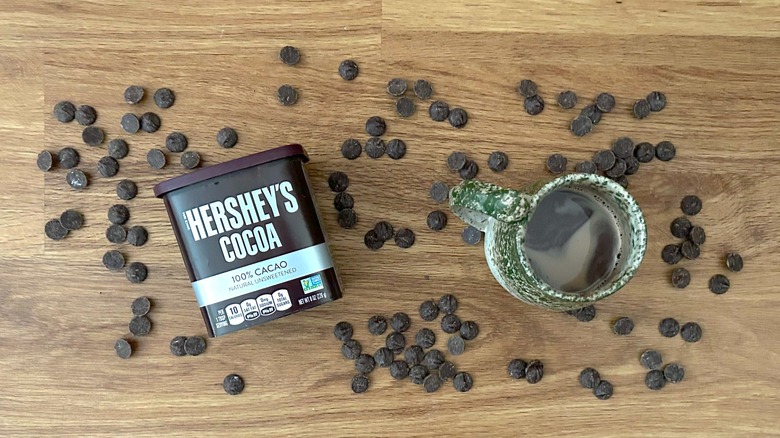 Container of Hershey's Cocoa powder and hot chocolate in a green mug surrounded by chocolate chips