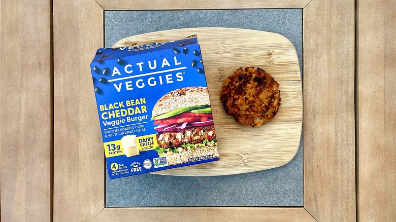A box of Actual Veggies Black Bean Cheddar Veggie Burgers on a cutting board next to a grilled patty