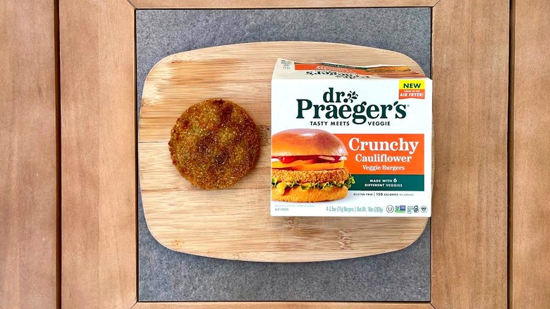 A grilled Dr. Praeger's Crunchy Cauliflower Veggie Burger on a cutting board next to a box of them