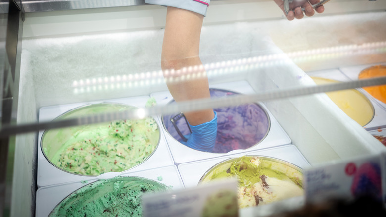 A person wearing a glove scooping ice cream from a display case with various colorful tubs of ice cream