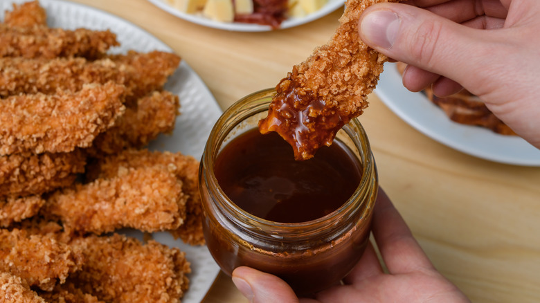 A hand dipping a chicken tender into BBQ sauce.