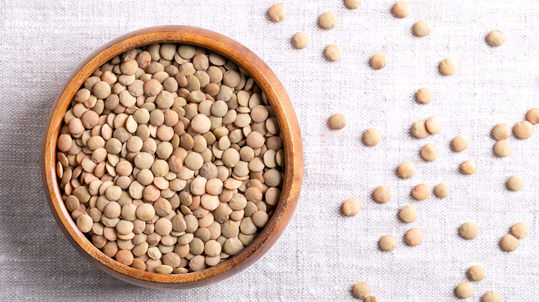 Pale brown lentils in a wooden bowl, bird's-eye view, with some lentils scattered across the surface to the right, all on a gray cloth background