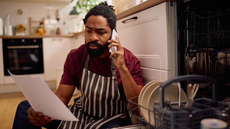 Man sitting on floor next to dishwasher while on phone and looking at document