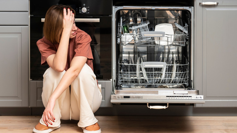 Unhappy woman sitting on floor next to open dishwasher