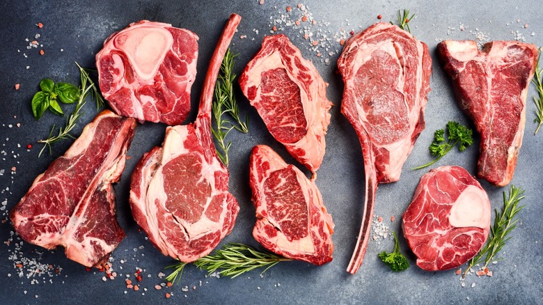 A variety of raw beef steaks with different seasonings and herbs around them.