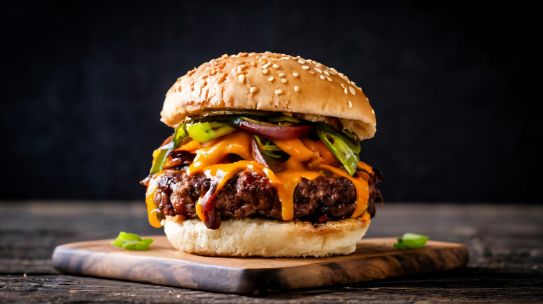 Cheeseburger with sesame seed bun and vegetable toppings on a small wooden cutting board