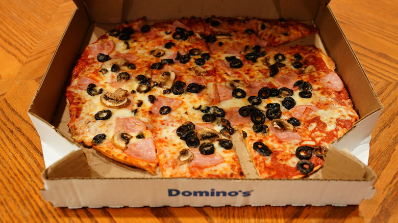 A square-cut Domino's pizza in a cardboard box on a wood table, topped with ham, black olives, and mushrooms, ready for consumption.