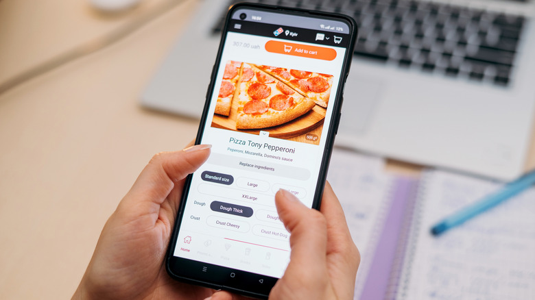 Hands holding a phone, ordering a pizza online through a food delivery app Domino's Pizza with a laptop and notebook visible on the desk.