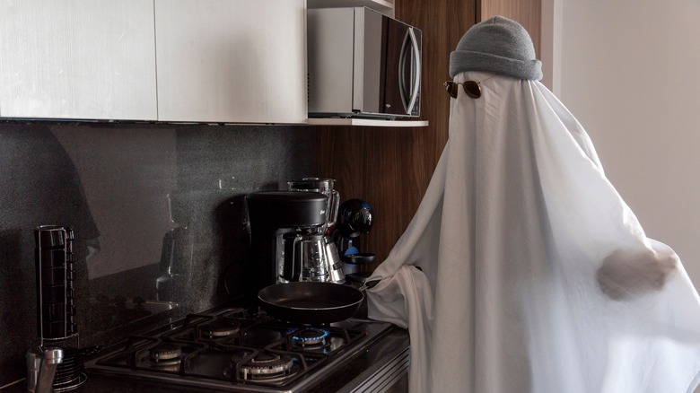 A person covered in a white sheet wearing sunglasses and a beanie while cooking at the stove