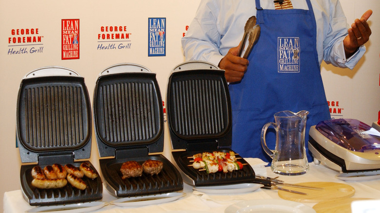 George Foreman standing next to 3 grills with meat on them.