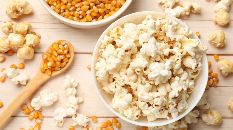 A bowl of popcorn surrounded by popcorn kernels on a table