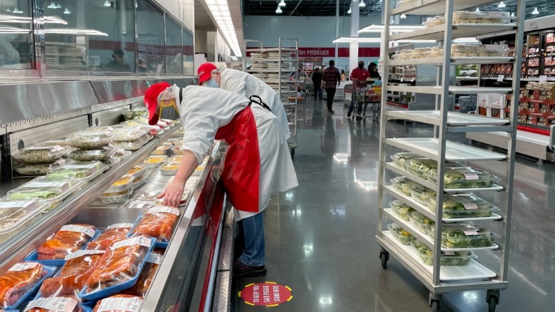 Two Costco employees restocking deli containers in a reach-in fridge inside of a warehouse store with a cart behind them.