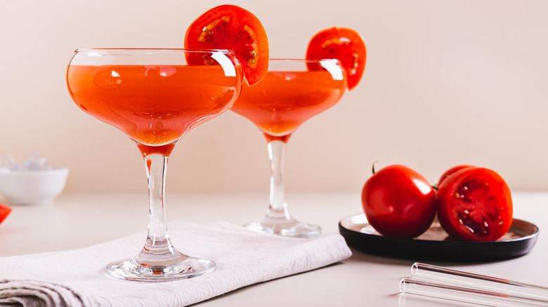 Two tomato martinis garnished with sliced tomatoes, alongside a small plate of tomatoes on a table