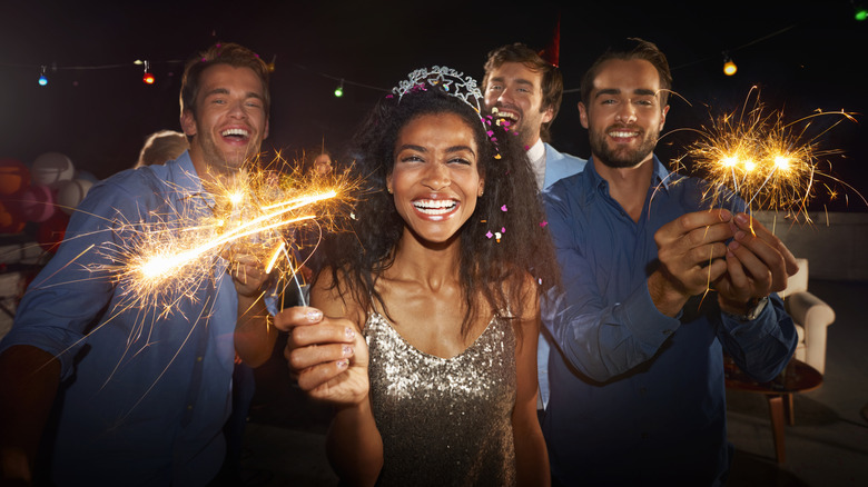 A group of people, including a woman wearing a glittery dress and a Happy New Year's crown, standing outside at night, smiling at the camera and holding sparklers