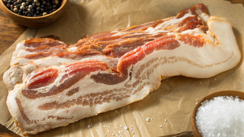 Thick cut bacon on brown paper with two small wood bowls of salt and peppercorns.