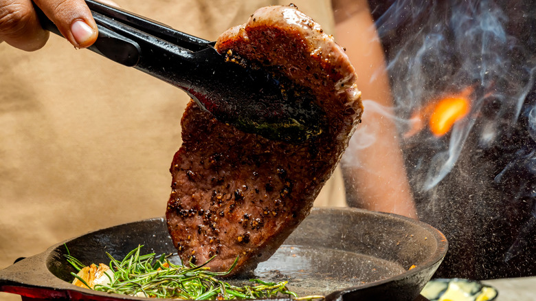 A person lifting a picanha steak out of a pan with tongs