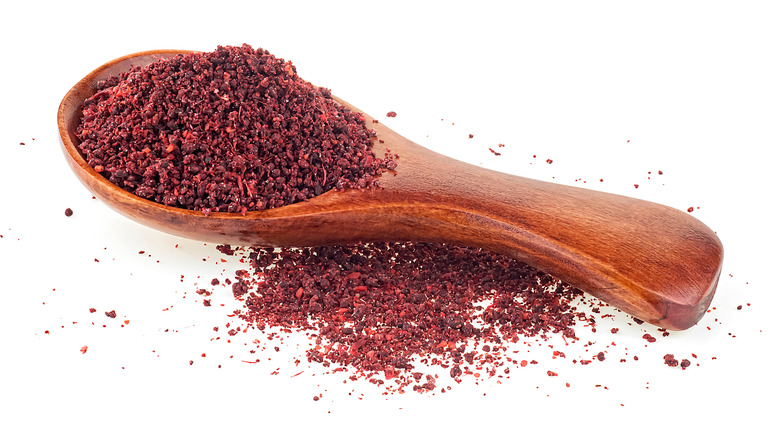Ground sumac in a wooden spoon with more sumac spilled near the handle, all on a white background